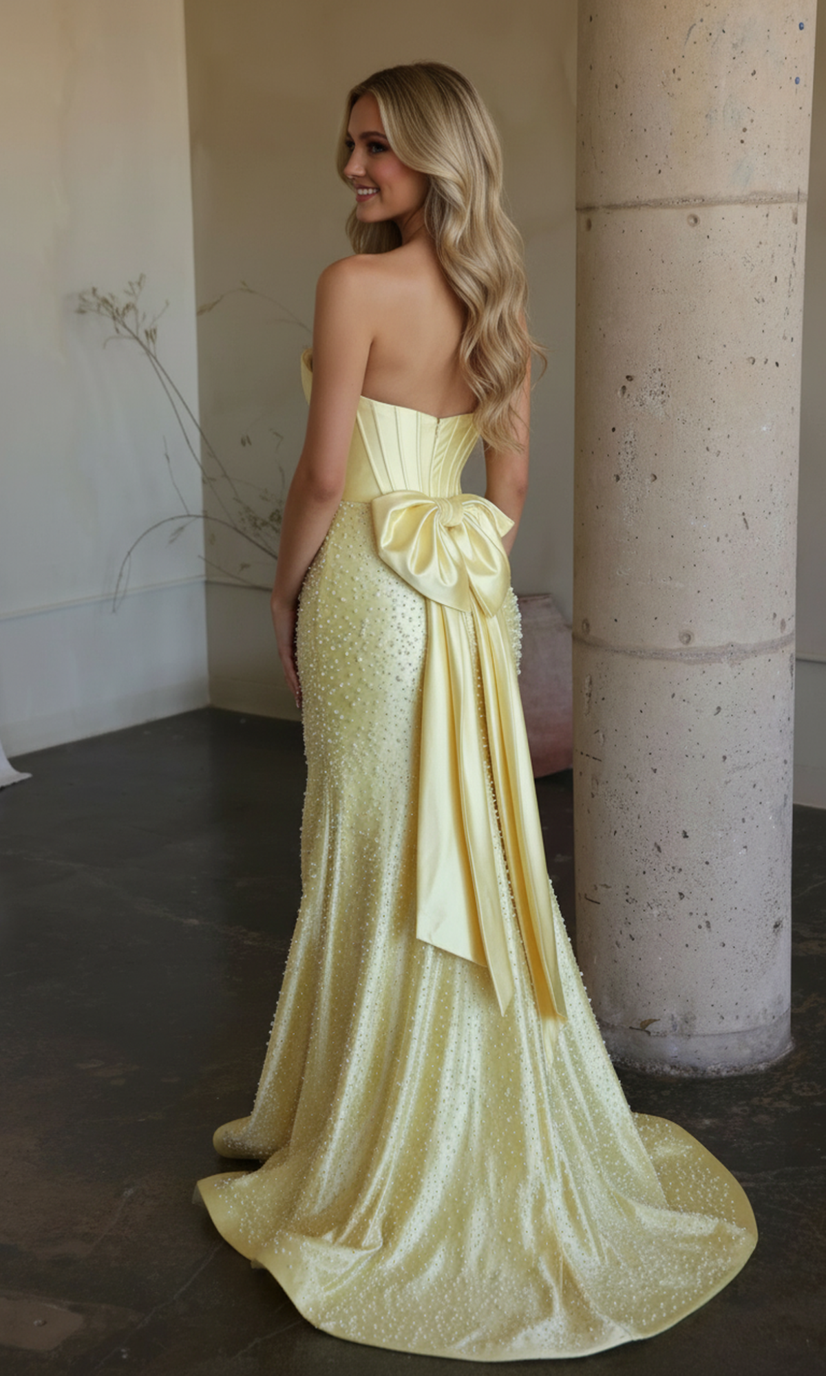 A woman wearing the Ava Presley 42513 long prom dress, featuring a strapless, pale yellow satin design with a large back bow, stands near a stone column, facing away and smiling.